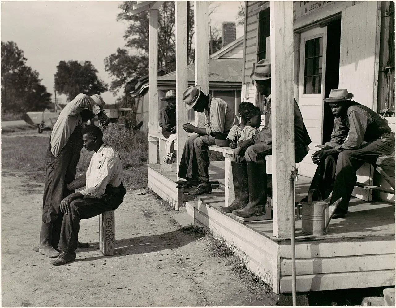 [Haircutting in Front of General Store and Post Office on Marcella Plantation, Mileston, Mississippi] by Marion Post Wolcott